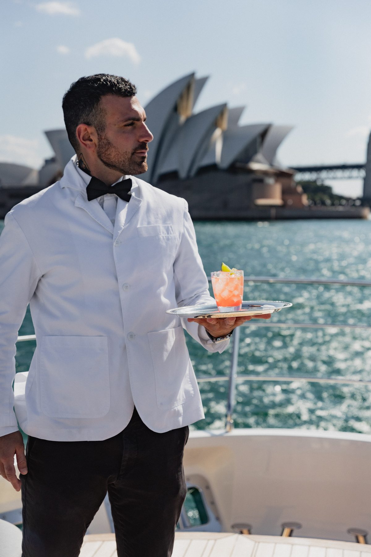 Icebergs waiter in a white jacket serving a cocktail on board Sirenuse, with the Sydney Opera House behind